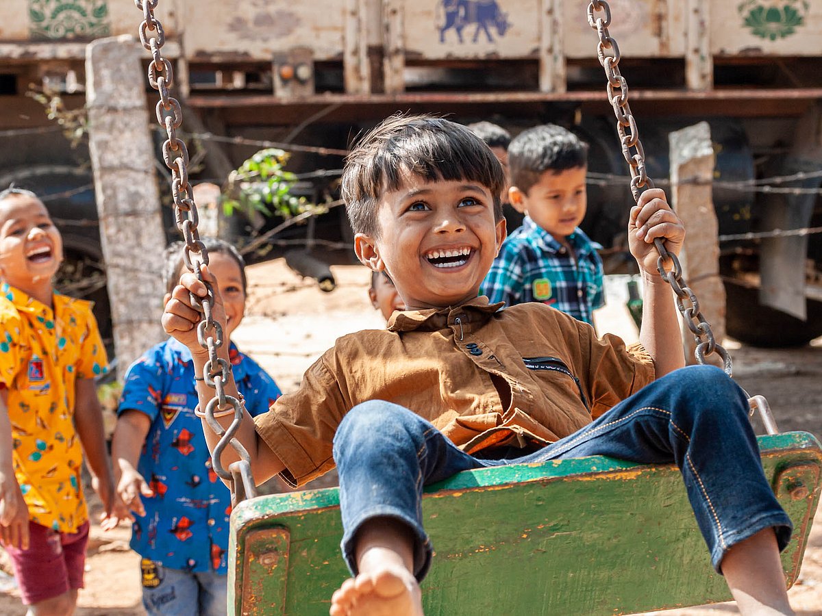 An Indian boy is swinging happily. In the background, a group of Indian children can be seen laughing.