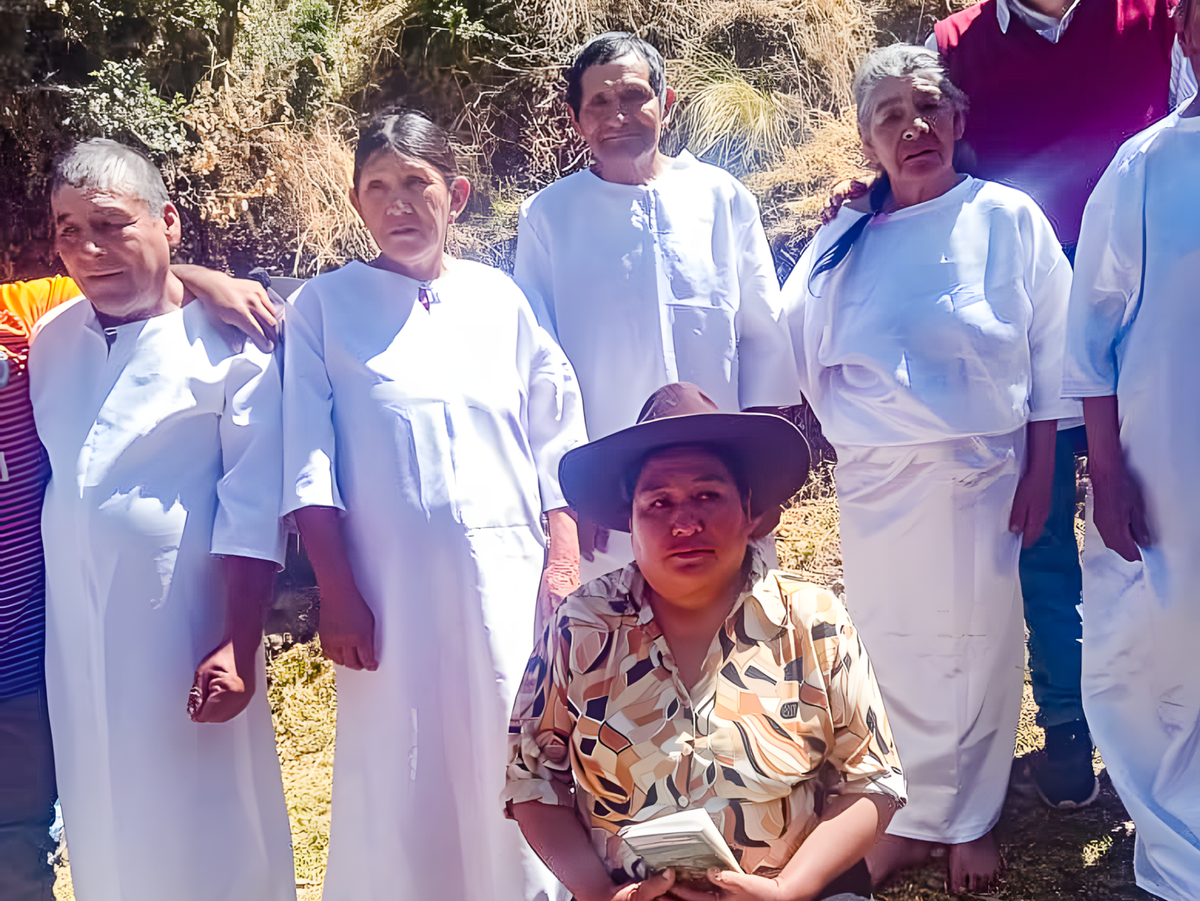 A Peruvian Quechua woman wearing a hat kneels facing the camera in front of a group of elderly people dressed in white, standing in a row behind her.