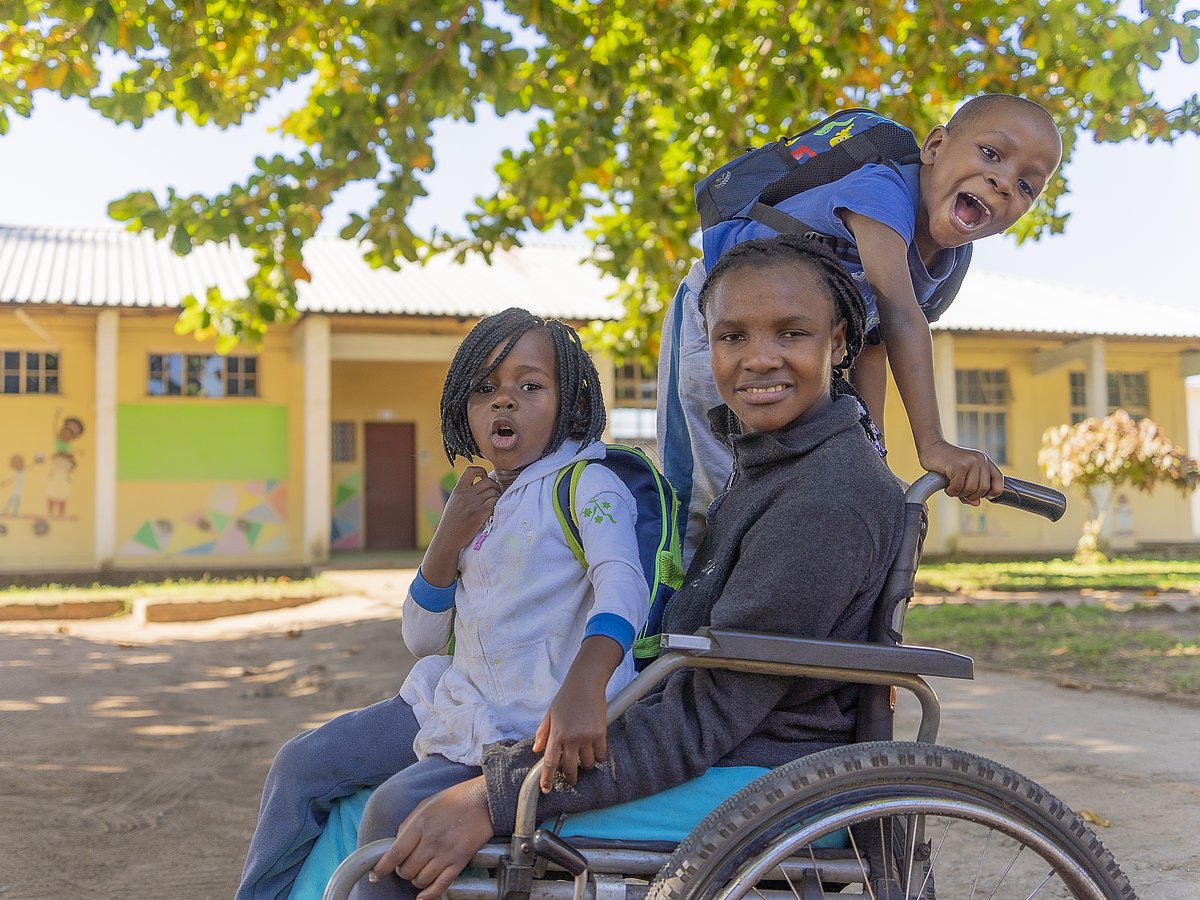 A young man from Mozambique in a wheelchair has a girl on his lap. A boy climbs onto the wheelchair and laughs cheekily.