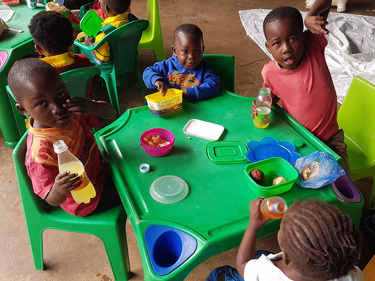 Eine Gruppe malawischer Kleinkinder sitzt um einen grünen Plastiktisch herum, der mit Tellern und Lebensmitteln gedeckt ist. Sie lächeln in die Kamera.