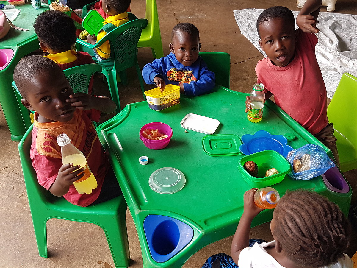 A group of Malawian preschool kids sit around a small plastic table laid with plastic plates, mugs and some food. They smile.