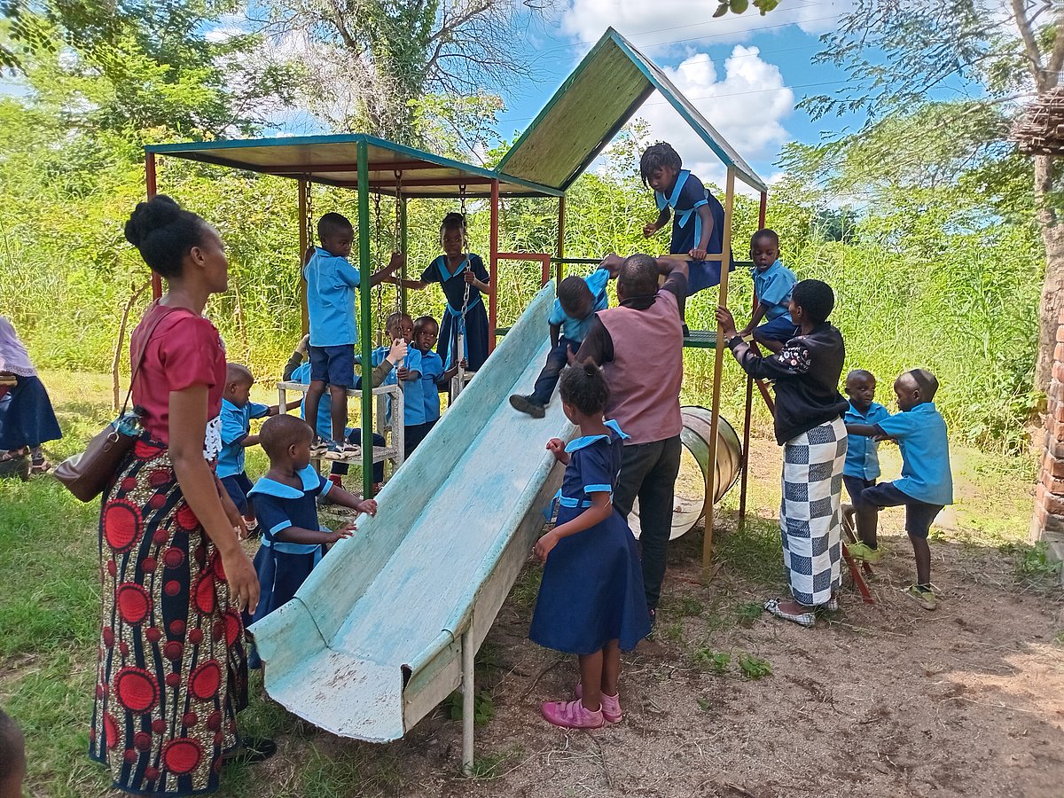 Ein Klettergerüst eines Spielplatzes mit einer Rutsche aus Metall wird umringt von einer Frau und Kindern in Malawi.
